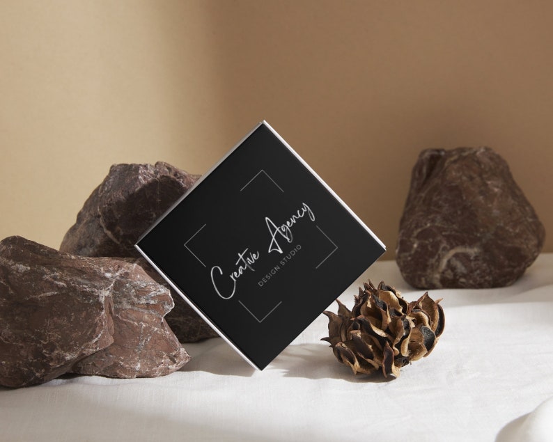 May include: A black square business card with silver lettering that reads "Creative Agency Design Studio" on a white background. The card is resting on a white surface with brown rocks and a dried flower.