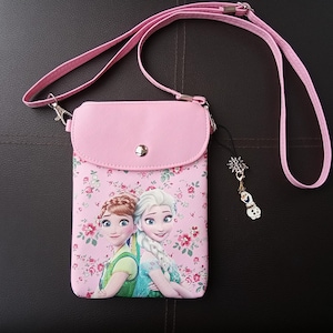 May include: A pink crossbody bag with a floral pattern and a cartoon image of two animated characters. The bag has a matching pink strap and a small charm with a snowflake and snowman design.