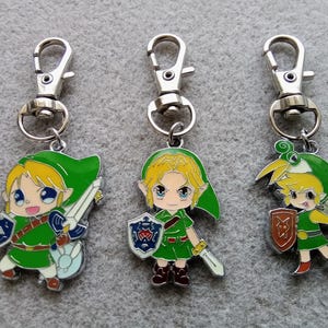 May include: Three silver keychains featuring cartoon characters from the Legend of Zelda video game series. Each keychain depicts a different character: Link, Zelda, and Ganondorf. The keychains are made of metal and have a green, gold, and red colour scheme.