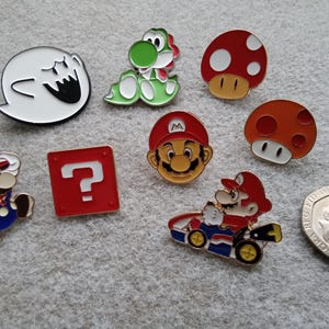 May include: A selection of enamel pins featuring characters from the Super Mario video game series. Includes Mario, Yoshi, mushrooms, a question block, and a Boo ghost. The pins are brightly coloured and detailed.