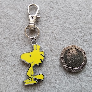 May include: A silver keychain with a yellow cartoon bird charm. The bird is standing with its wings down and has a yellow beak and black eyes.
