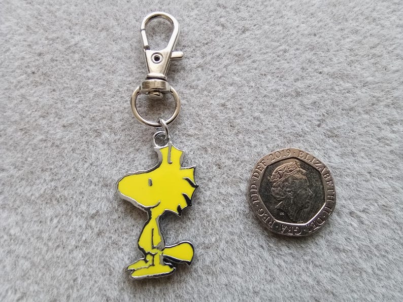 May include: A silver keychain with a yellow cartoon bird charm. The bird is standing with its wings down and has a yellow beak and black eyes.