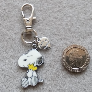 May include: Silver keychain featuring a cartoon Snoopy holding a yellow Woodstock bird charm. The keychain also has a heart-shaped charm with the text "Best Friend" on it.
