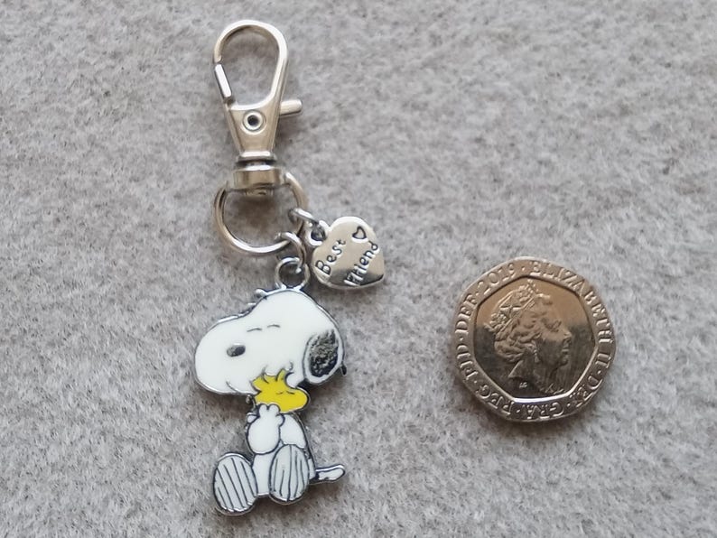 May include: Silver keychain featuring a cartoon Snoopy holding a yellow Woodstock bird charm. The keychain also has a heart-shaped charm with the text "Best Friend" on it.