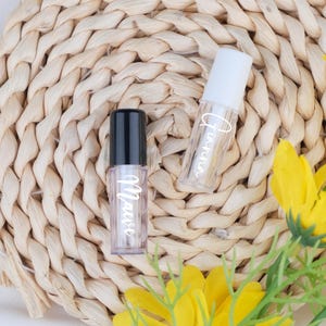 Op de afbeelding: Twee doorzichtige lipgloss tubes met witte tekst, één met de tekst "Maese" en de andere met "Tianna". De tubes hebben zwarte en witte doppen. De achtergrond is een geweven stro mat met gele bloemen.
