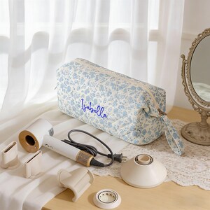 May include: A white and gold hair dryer with multiple attachments and a floral print cosmetic bag with the name "Isabella" embroidered in blue. The bag and hair dryer are on a wooden surface.