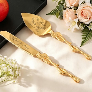May include: A gold cake server and knife set. The server has a wide, triangular blade, and the knife has a serrated edge. Both feature ornate floral handles and the engraved names "Arthur & Freya" and "The Smith" with the date "EST.2026".