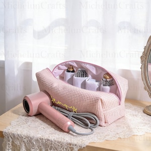 May include: A pink waffle-textured cosmetic bag with a zipper, containing hair care products. A pink hairdryer and a gray cord are in front of the bag. The bag has the word "Jenny" embroidered in yellow.