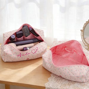May include: Two pink floral quilted cosmetic bags. One bag is open, revealing various hair styling tools and brushes. The interior lining is a pink and white gingham pattern. The other bag is also open, showing the interior pockets. The text "Amelia Love" is embroidered on the bag.