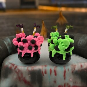 May include: Two black cauldron-shaped containers with pink and green candy skulls and sprinkles. The containers are decorated with black and white striped straws.