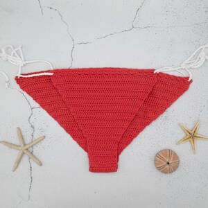 May include: A red crocheted bikini bottom with white ties. The bikini bottom is lying on a white textured surface.