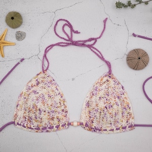Pode incluir: Um top de biquíni de crochê com um padrão floral roxo e branco. O top tem alças ajustáveis e um fecho de nó nas costas.
