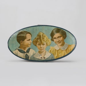 May include: Oval-shaped tin with a blue background and a picture of three children. The children are wearing different outfits and have different hairstyles. The tin is likely for storing small items.