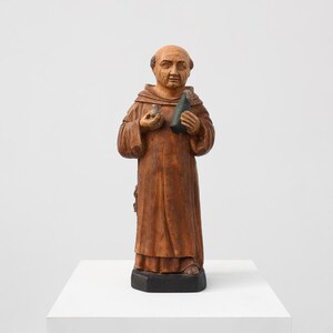 Könnte beinhalten: Holzstatue eines Mönchs in brauner Robe, der eine kleine Flasche und eine Phiole hält. Die Statue hat eine kahle Kopf und steht auf einem schwarzen Sockel. Die Figur ist detailliert geschnitzt.