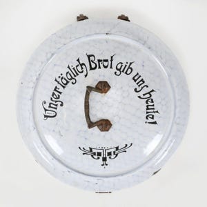 Peut inclure: Couvercle en émail blanc avec un motif moucheté et une poignée sombre. Le couvercle porte l'inscription allemande "Unser täglich Brot gib uns heute!" en noir. Des volutes décoratives noires sont en dessous.