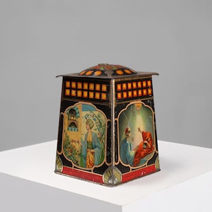 Pode incluir: Uma caixa de lata vintage com fundo preto e ilustrações coloridas de pessoas com roupas tradicionais. A caixa tem formato quadrado com uma tampa abobadada e uma borda decorativa. O texto na caixa diz "Chocolat de Luxe" e "Fabriqué en Suisse".