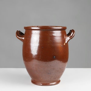 Op de afbeelding: Een bruine aardewerken pot met twee lusvormige handvatten. De pot heeft een rond lichaam en een licht uitlopende lip.