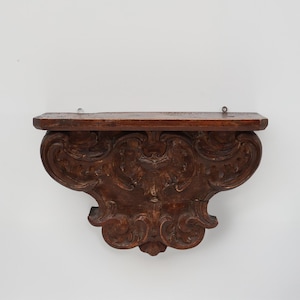 May include: A dark brown, antique wooden wall shelf with ornate, carved details. The shelf has a rectangular top and a decorative, scroll-like bracket. Two metal rings are attached to the top for hanging. The shelf is likely made of solid wood.