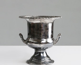 Puede incluir: Una urna de metal plateado con un borde ancho y texturizado y dos asas curvas. La urna tiene un cuerpo bulboso y una base de pedestal. La superficie refleja la luz, creando una apariencia brillante. El fondo es gris liso.