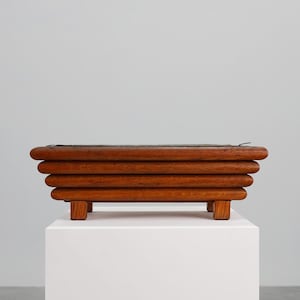 Peut inclure: Un bac à plantes rectangulaire en bois avec une finition foncée. La boîte est construite à partir de lattes de bois horizontales et arrondies et repose sur quatre petits pieds carrés. L'intérieur semble être doublé d'un matériau sombre. Le pot est exposé sur un piédestal blanc.