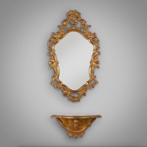 Könnte beinhalten: Ein verzierter goldfarbener Spiegel mit einem dekorativen Regal darunter. Der Spiegel hat eine gewellte Form und komplizierte Details.