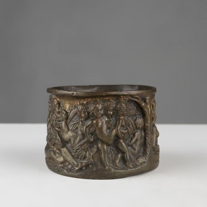 Op de afbeelding: Een ovale bronzen plantenbak met een reliëf van figuren en bomen aan de zijkanten.