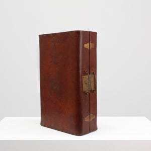 May include: A vintage, rectangular, brown leather case with brass hardware. The case has a hinged closure with a brass latch and handle. Brass corner accents add to the antique aesthetic. The case is standing upright on a white surface.