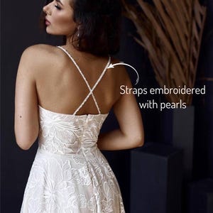 A woman wearing a white lace dress with straps embroidered with pearls, posing against a dark background.