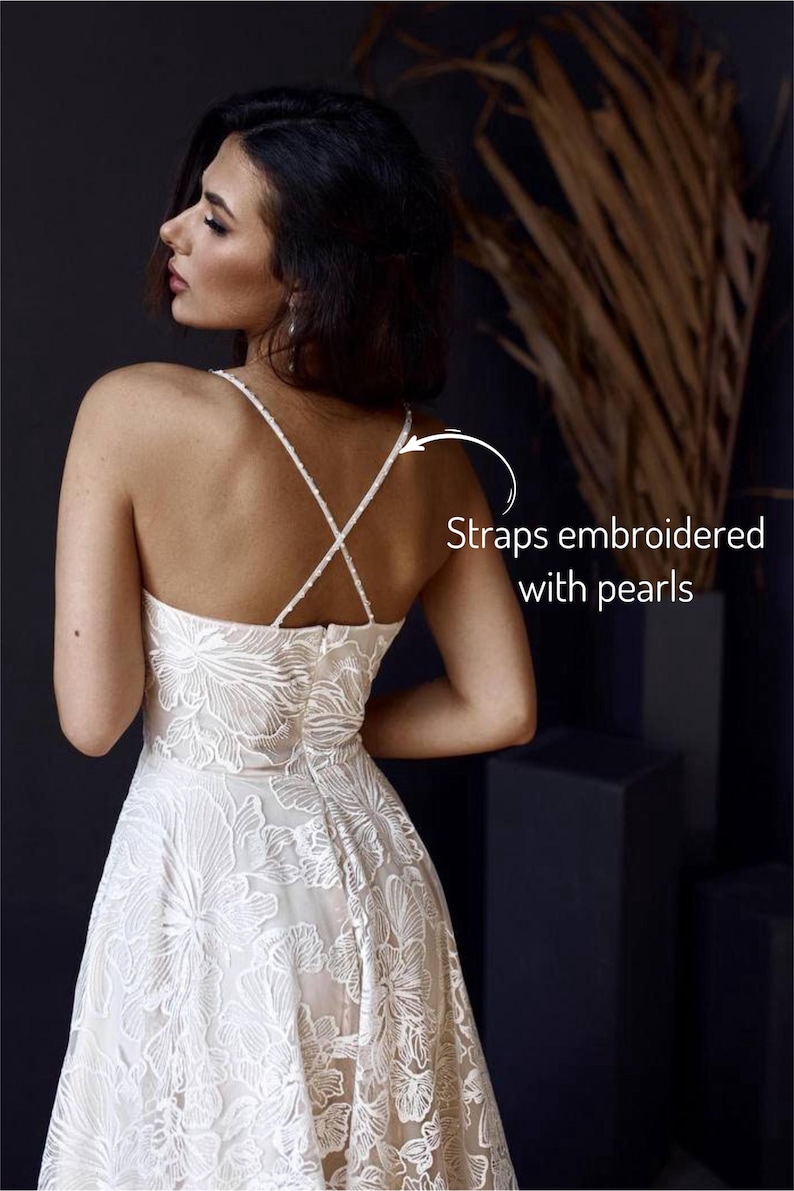 A woman wearing a white lace dress with straps embroidered with pearls, posing against a dark background.