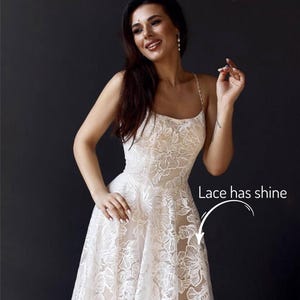 A woman in a white lace dress with a floral pattern, smiling and posing against a dark background.