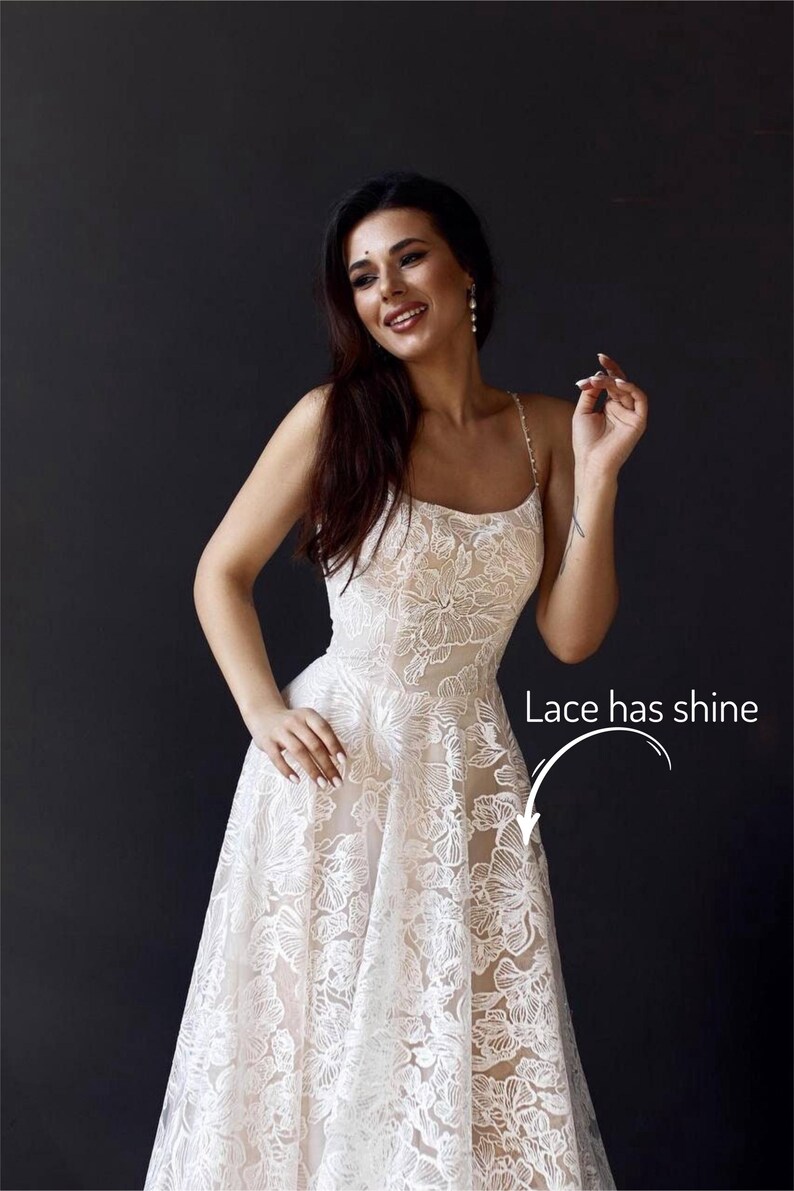 A woman in a white lace dress with a floral pattern, smiling and posing against a dark background.