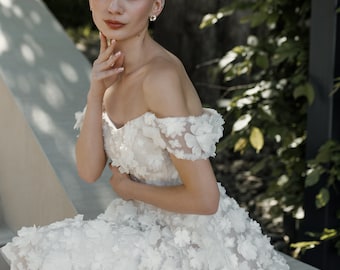 May include: A woman wearing a white, off-the-shoulder dress adorned with three-dimensional floral appliqués. The dress features a sweetheart neckline and a knee-length skirt. The woman is seated outdoors, with greenery in the background.