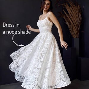 A woman in a white lace dress with a flowing skirt is posing in front of a dark background, with a plant and a text overlay that reads "Dress in a nude shade".