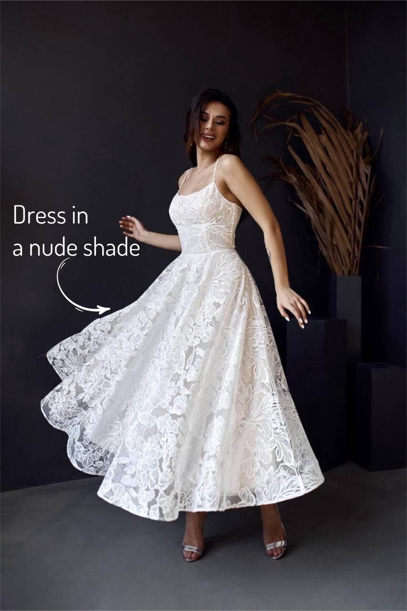 A woman in a white lace dress with a flowing skirt is posing in front of a dark background, with a plant and a text overlay that reads "Dress in a nude shade".