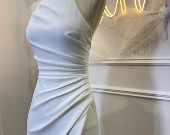 May include: A white sleeveless dress featuring a high neckline and a fitted bodice with diagonal ruching details along the side. The garment is displayed on a mannequin, showcasing a smooth fabric texture and a structured silhouette that extends downward.