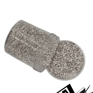 May include: A clear plastic container filled with small, light gray gravel. The container is tipped over, spilling the gravel onto a white surface. The container has a label that reads "ROCK-N-WOOD | SUPPLY YARD".