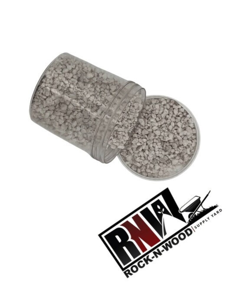 May include: A clear plastic container filled with small, light gray gravel. The container is tipped over, spilling the gravel onto a white surface. The container has a label that reads "ROCK-N-WOOD | SUPPLY YARD".