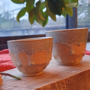 Peut inclure: Deux tasses à thé en céramique avec un glaçage moucheté dans les tons gris, blanc et orange. Les tasses ont un effet de glaçage coulant, ce qui leur donne un aspect unique et rustique.