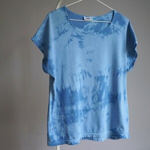 Peut inclure: Un t-shirt bleu clair tie-dye à manches courtes est suspendu à un cintre blanc. Le t-shirt a un col rond et un aspect délavé et texturé. Le motif tie-dye est d'une nuance de bleu plus foncée.