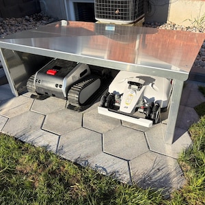 May include: A silver metal shelter houses two robotic lawn mowers. One is black and gray with tank treads and a red button. The other is white with black wheels and a yellow emblem. The shelter sits on gray hexagonal pavers.