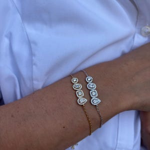 Peut inclure: Deux bracelets délicats, l'un doré et l'autre argenté, chacun présentant une rangée centrale de trois pierres claires aux formes distinctes : une ronde, une carrée et une en forme de poire, entourées d'un halo de petites pierres scintillantes.