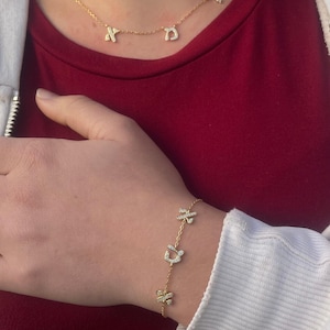 Peut inclure: Un collier doré avec trois petits pendentifs de lettres scintillantes. Un bracelet doré assorti avec trois breloques décoratives est également visible. Les bijoux sont mis en valeur sur un haut rouge et une veste blanche.