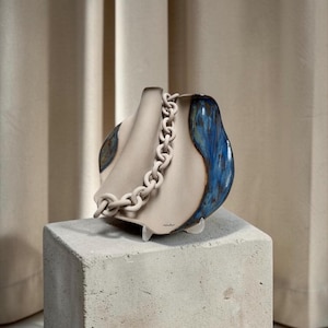 Op de afbeelding: Een keramische vaas met een unieke vorm, met een crèmekleurig lichaam met blauwe geglazuurde accenten en een crèmekleurige ketting als handvat. De vaas staat op een betonnen blok, wat het moderne ontwerp benadrukt.