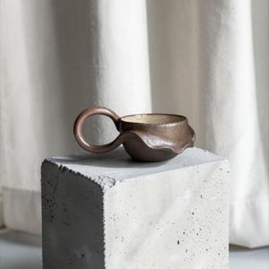Op de afbeelding: Een handgemaakte bruine keramische mok met een uniek, golvend ontwerp en een ronde handgreep. De binnenkant heeft een lichtere, gespikkelde afwerking. De mok staat op een grijze betonnen blok tegen een witte achtergrond.