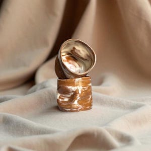 Op de afbeelding: Twee handgemaakte keramische bekers op elkaar gestapeld. De bekers hebben een bruin en wit gemarmerd ontwerp. De bovenste beker is gekanteld en onthult de binnenkant met een vergelijkbaar kleurenschema. De bekers staan tegen een neutrale achtergrond.