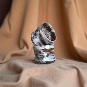 Op de afbeelding: Twee handgemaakte keramische bekers die op elkaar zijn gestapeld. De bekers hebben een unieke, organische vorm en zijn geglazuurd met een mix van bruine, witte en blauwe tinten. Het getextureerde oppervlak draagt bij aan hun artistieke aantrekkingskracht.