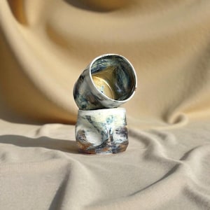 Op de afbeelding: Twee handgemaakte keramische bekers op elkaar gestapeld. De onderste beker is wit met bruine en blauwe accenten. De bovenste beker is gekanteld en onthult de binnenkant. De bekers hebben een rustieke, getextureerde uitstraling.