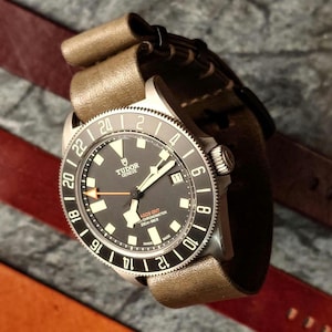 Può includere: Un orologio Tudor nero e argento con un cinturino in pelle marrone. Il quadrante dell'orologio ha uno sfondo nero con marcature bianche e una lancetta dei secondi rossa. L'orologio ha una finestrella della data alle 3 e una ghiera girevole con numeri e marcature.