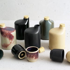 Puede incluir: Un conjunto de botellas de cerámica con tapones de corcho en varios colores y patrones, incluyendo negro, gris, beige y un gris moteado y morado. También hay varias tazas pequeñas de cerámica en negro, beige y gris moteado y morado.