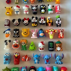 May include: A collection of small, colorful PVC character charms featuring various pop culture figures, including superheroes, cartoon animals, movie characters, and beverage cans, arranged in a grid on a plain white background.